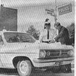 Two police officers lean over the bonnet of a late 1960's model Ford Falcon police car