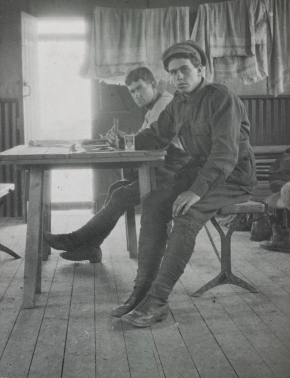 Two men, whole-length, to right, man in foreground wearing uniform with peaked cap and puttees, man behind with jacket off, also wearing puttees, both seated at a wooden table, towels draped over line and open door in background.